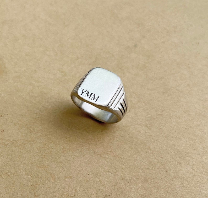May include: A silver signet ring with a square top and a grooved band. The top of the ring is engraved with the letters "YMMI".