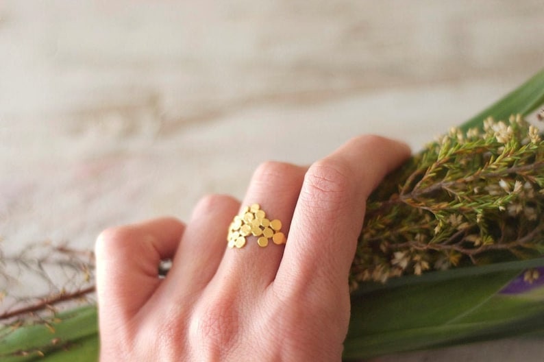 May include: A gold ring with a unique design featuring a cluster of small, round, gold discs. The ring is worn on a finger and the hand is holding a bouquet of green foliage.