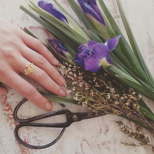 May include: A gold ring with a delicate design of small circles, worn on a hand resting on a bouquet of purple irises and green foliage.