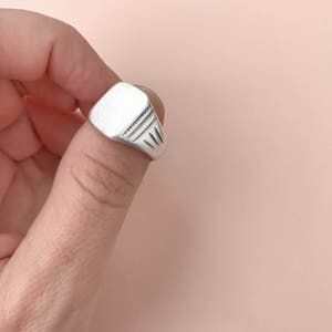 May include: A silver signet ring with a smooth, flat top and a textured band.