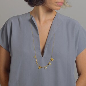 May include: A gold necklace with a geometric design featuring circles, triangles, and a small cluster of beads. The necklace is worn by a person with short, dark hair and a gray shirt.