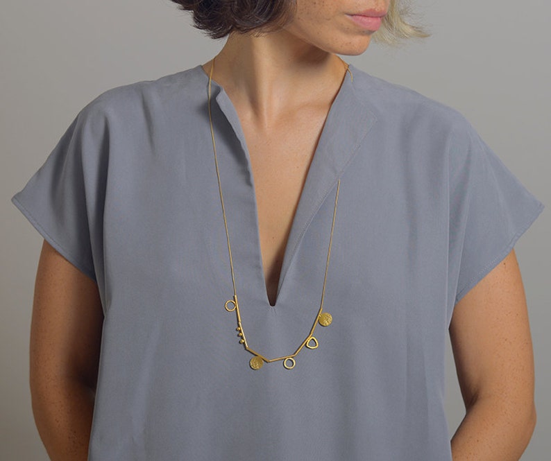 May include: A gold necklace with a geometric design featuring circles, triangles, and a small cluster of beads. The necklace is worn by a person with short, dark hair and a gray shirt.