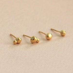 Może przedstawiać: Cztery pary złotych kolczyków. Dwie pary mają pojedynczy okrągły kolczyk, a pozostałe dwie pary mają dwa okrągłe kolczyki w różnych rozmiarach.