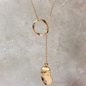 Puede incluir: Un collar de oro con un colgante de círculo retorcido y un colgante con forma de cara.