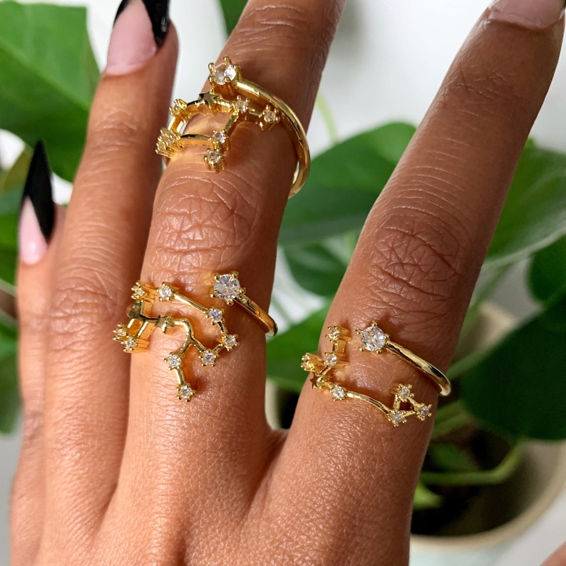 May include: Three gold rings with constellation designs and clear rhinestones. The rings are worn on the fingers of a hand.