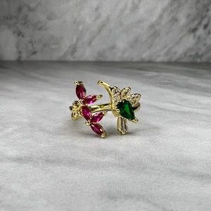 May include: Gold ring with a hummingbird design. The hummingbird has green and clear rhinestones. The ring also has a branch with pink rhinestones.