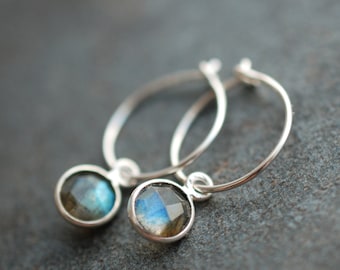 May include: A pair of silver hoop earrings featuring round, faceted labradorite gemstones. Each stone is set in a simple silver bezel, suspended from a thin, circular silver wire hoop. The labradorite stones display iridescent blue flashes.