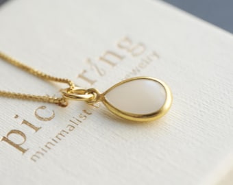 May include: A gold chain necklace with a teardrop-shaped pendant featuring a smooth white stone inlay. The necklace rests on a white square box with the text "picring minimalist jewelry" printed in gold lettering on the top surface.