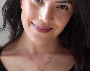 May include: A delicate gold necklace featuring two small, interlocking circular rings as a pendant. The thin gold chain rests against a black sleeveless top. The design is simple and consists of a polished metallic finish that catches the light.