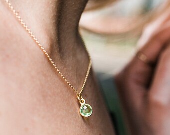 May include: A delicate gold chain necklace featuring a single, round, faceted light green gemstone pendant set in a simple gold bezel setting. The necklace rests against the skin, showcasing the reflective quality of the metal and the stone.
