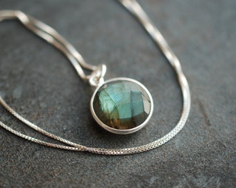 May include: A round, faceted labradorite gemstone pendant set in a polished silver bezel. The stone displays iridescent blue and green flashes. The pendant hangs from a delicate silver box chain necklace, creating a simple and elegant piece of jewelry.