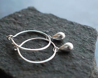 May include: Silver hoop earrings with teardrop pendants. The earrings are made of a shiny, reflective metal and are resting on a dark gray, textured surface. The teardrop pendants hang from the bottom of the hoops.