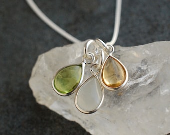 May include: A silver necklace featuring three teardrop-shaped pendants. The pendants are filled with green, white, and yellow gemstones, each with a silver border. The necklace is displayed on a white, textured surface.