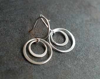 May include: Silver-toned earrings with a nested circle design. Each earring features three concentric circles of varying sizes, suspended from a simple hook. The jewelry is displayed against a dark, textured background.