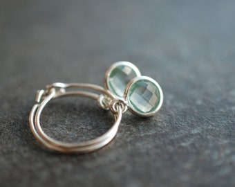 May include: Silver hoop earrings with a light green gemstone dangle. The earrings are made of a thin silver wire and have a simple design.