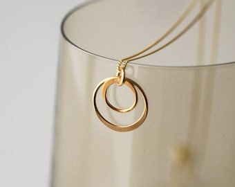 May include: A gold-toned necklace featuring a pendant composed of two interlocking circular rings of different sizes. The pendant is suspended from a delicate, thin gold-toned chain, creating a simple and clean design with a polished metallic finish.
