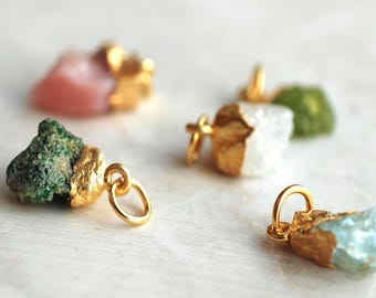 May include: Four raw gemstone pendants featuring irregular, unpolished stones in shades of pink, dark green, white, and light blue. Each stone is partially dipped or capped in a textured, metallic gold-tone finish and includes a small gold jump ring.