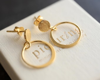 May include: A pair of gold-toned stud earrings featuring a small circular disc attached to a larger open hoop. The earrings are resting on a white textured box with the text "piourins" embossed on the surface, showing a metallic finish.