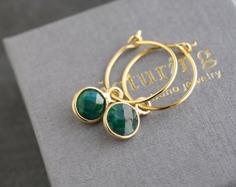 May include: A pair of gold hoop earrings featuring round, faceted dark green gemstone charms. The earrings are crafted with thin gold wire hoops and circular settings that hold the polished green stones, creating a simple and elegant jewelry piece.