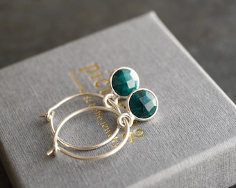 May include: A pair of gold hoop earrings with round, faceted, emerald-green gemstones. The earrings rest on a gray textured box. The earrings are a jewelry item.
