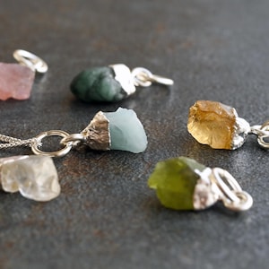 Könnte beinhalten: Eine Sammlung von Edelsteinanhängern in verschiedenen Farben, darunter Rosa, Grün, Blau und Gelb. Jeder Anhänger hat einen rohen, ungeschliffenen Stein, der in einem silberfarbenen Metall gefasst ist. Die Anhänger liegen auf einer dunkelgrauen Oberfläche.