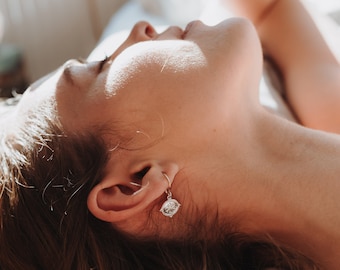 May include: A close-up of a person's face with a silver earring with a clear stone pendant. The person is lying down with their eyes closed.