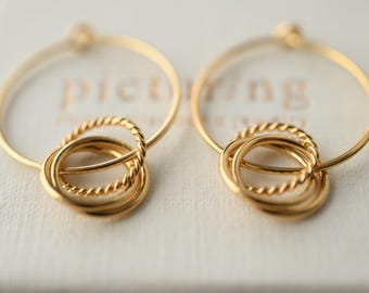 May include: A pair of gold-colored hoop earrings. Each earring features a large, smooth circular hoop with a smaller, textured, intertwined design at the bottom. The earrings are displayed on a white surface.