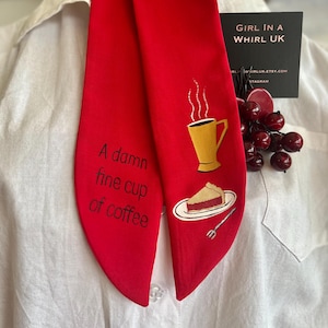 May include: A vibrant red necktie featuring the phrase "A damn fine cup of coffee" and a graphic of a yellow coffee cup and cake slice. The tie is displayed on a white shirt.