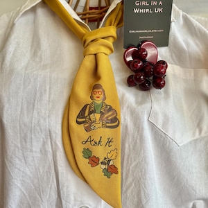 May include: Mustard yellow necktie featuring a cartoon figure holding a drink, the text "Ask It", and autumn leaves. A red heart-shaped brooch with red beads and a business card are also visible.