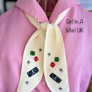 May include: A pink shirt with a cream-coloured scarf tied at the neck. The scarf features dice and domino designs, and starburst accents. The text "Girl In A Whirl UK" is visible on the right side of the image.