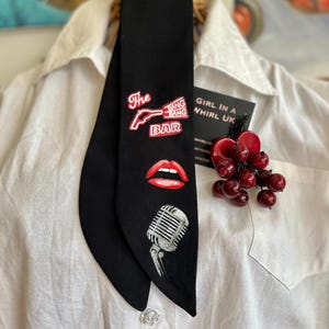 May include: Black necktie featuring red and white graphics: a gun, the words "The Bang Bang Bar", red lips, and a vintage microphone. The tie is displayed on a white collared shirt.
