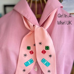 May include: A pale pink shirt with a matching scarf tied in a bow. The scarf features dice and domino designs, and the text "Girl In A Whirl UK" is printed on the shirt.