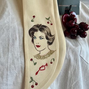 May include: A cream-coloured scarf with an embroidered design of a woman's face and cherries. The scarf has a vintage style, with the text "GIRL IN A WHIRL UK" on a tag. A cherry-shaped decoration is also visible.