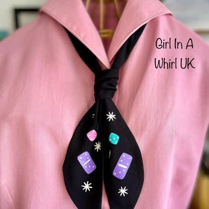 May include: A black necktie with a domino and star pattern, knotted, against a pink collared shirt. The tie features white stars, pink, teal, and purple dominoes. The text "Girl In A Whirl UK" is visible.