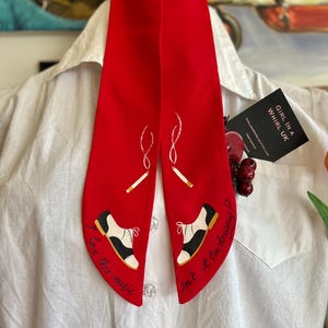 May include: A red necktie with a decorative design. The tie features a pair of black and white shoes with gold accents, cigarettes, and the phrases "I love this music" and "Isn't it too dreamy?" The tie is displayed on a white shirt.