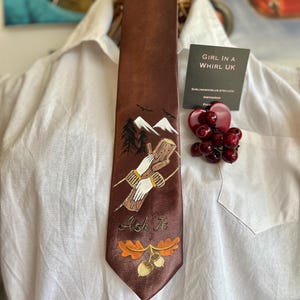 May include: A brown tie with an embroidered design featuring mountains, trees, and hands holding a log. The tie has the text "Ask It" and oak leaf and acorn details. The tie is displayed on a white shirt.