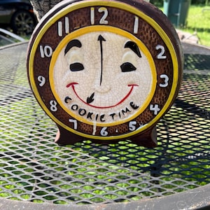 Puede incluir: Un reloj de cerámica con una cara sonriente y las palabras "COOKIE TIME" en la esfera. El reloj tiene un marco marrón con un borde amarillo y números blancos. Las manecillas señalan las 12 en punto.