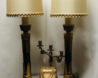 May include: Two gold and black table lamps with cream-colored lampshades trimmed in gold. The lamps are on a white table with a gold decorative box in the center.