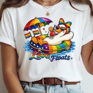 Pode incluir: Uma t-shirt branca com o gráfico de um pato branco a usar óculos de sol arco-íris e um laço, a relaxar numa boia arco-íris sob um guarda-sol arco-íris. O pato segura um cocktail e o texto Love Floats está impresso na parte inferior.