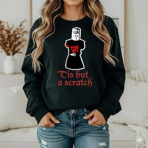 May include: Black sweatshirt featuring a graphic of a knight in armor with a red torso and the text "'Tis but a scratch" in red. The design is centered on the front of the long-sleeved top.