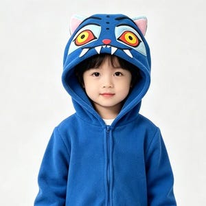 May include: A blue fleece onesie featuring a hood designed to look like a creature with large yellow eyes, pointed ears, and white teeth. The garment has long sleeves, a front zipper closure, and ribbed cuffs at the wrists and ankles.