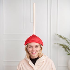 Op de afbeelding: Een vrouw draagt ​​een rode zuigerhoed met een witte stok die van de bovenkant uitsteekt. De hoed heeft een wit elastiek dat om het hoofd gaat.