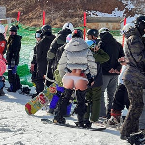 Pode incluir: Um grupo de snowboarders em roupas de inverno, incluindo capacetes e jaquetas, em pé em uma encosta nevada. Um snowboarder tem um gráfico engraçado em suas calças. Uma prancha de snowboard com um design floral colorido é visível.