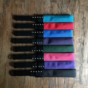 May include: Nine black leather straps with different colored fabric inserts. The straps have silver studs and are lined up vertically. The colors of the fabric inserts are turquoise, red, blue, purple, green, pink, purple, and black.