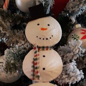 Op de afbeelding: Een sneeuwman ornament gemaakt van drie witte ronde zanddollars met een zwarte hoed, zwarte ogen, een kleine oranje neus en een rood-witte geruite sjaal.