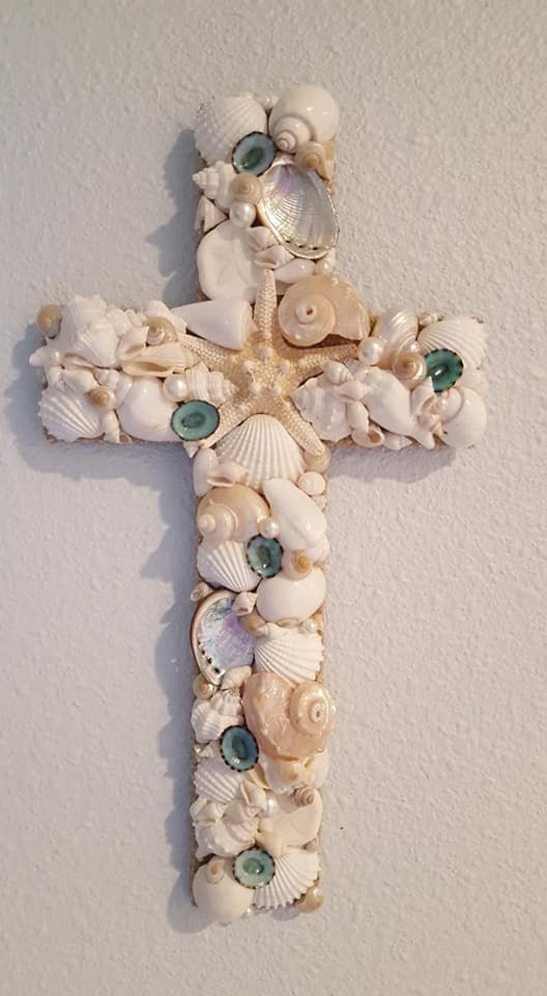 May include: A white wooden cross decorated with seashells, starfish, and pearls. The cross is made of natural materials and has a rustic, coastal feel.