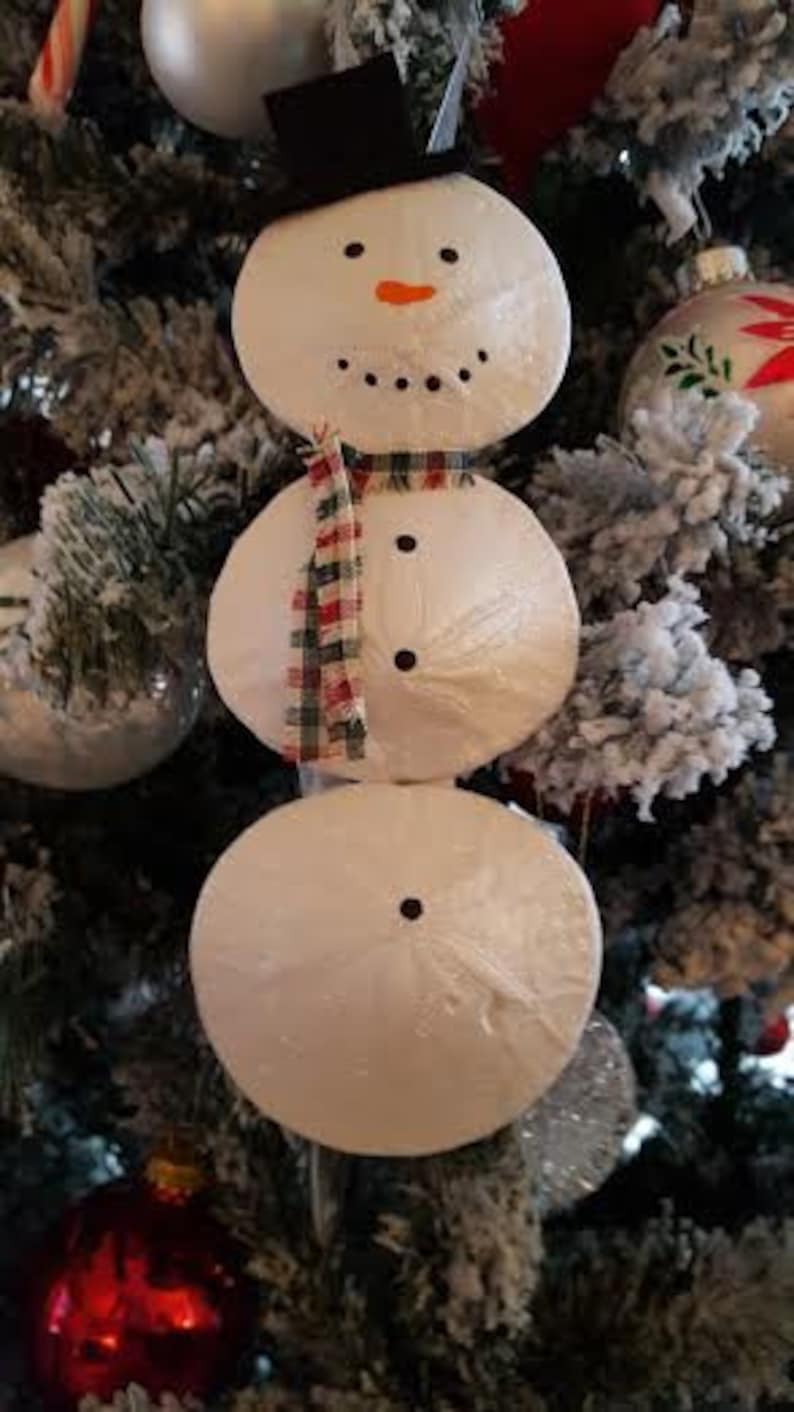 Op de afbeelding: Een sneeuwman ornament gemaakt van drie witte ronde zanddollars met een zwarte hoed, zwarte ogen, een kleine oranje neus en een rood-witte geruite sjaal.