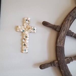 May include: A white seashell cross with green glass accents hangs on a white wall next to a brown wooden ship's wheel.