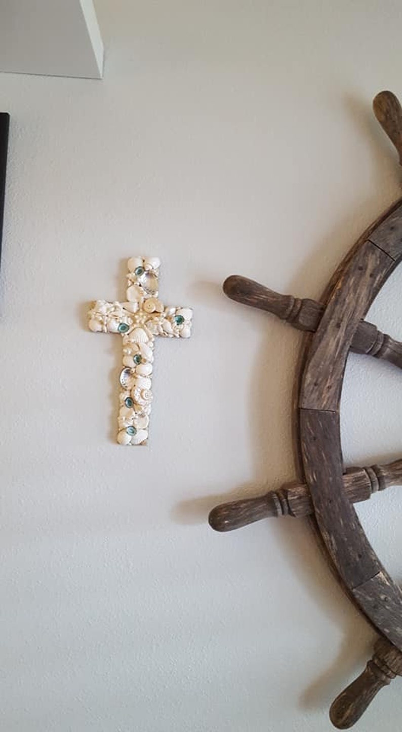 May include: A white seashell cross with green glass accents hangs on a white wall next to a brown wooden ship's wheel.