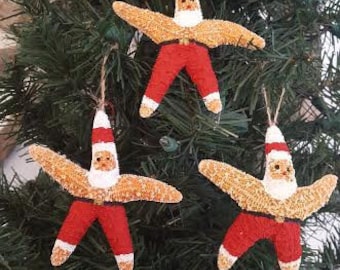 May include: Three Christmas tree ornaments shaped like starfish, each adorned with a Santa Claus design. The ornaments feature a tan starfish body, red pants, a white beard, and a Santa hat. They hang from a green Christmas tree.